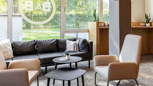 A modern living room with a grey sofa, two round tables, and a beige armchair. A large window offers a view of greenery outside. - B&B Hotel Hamburg-Nord (Hamburg)