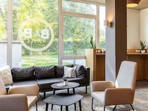 Un salon moderne avec un canapé gris, deux tables rondes et un fauteuil beige. Une grande fenêtre offre une vue sur la verdure à l’extérieur.