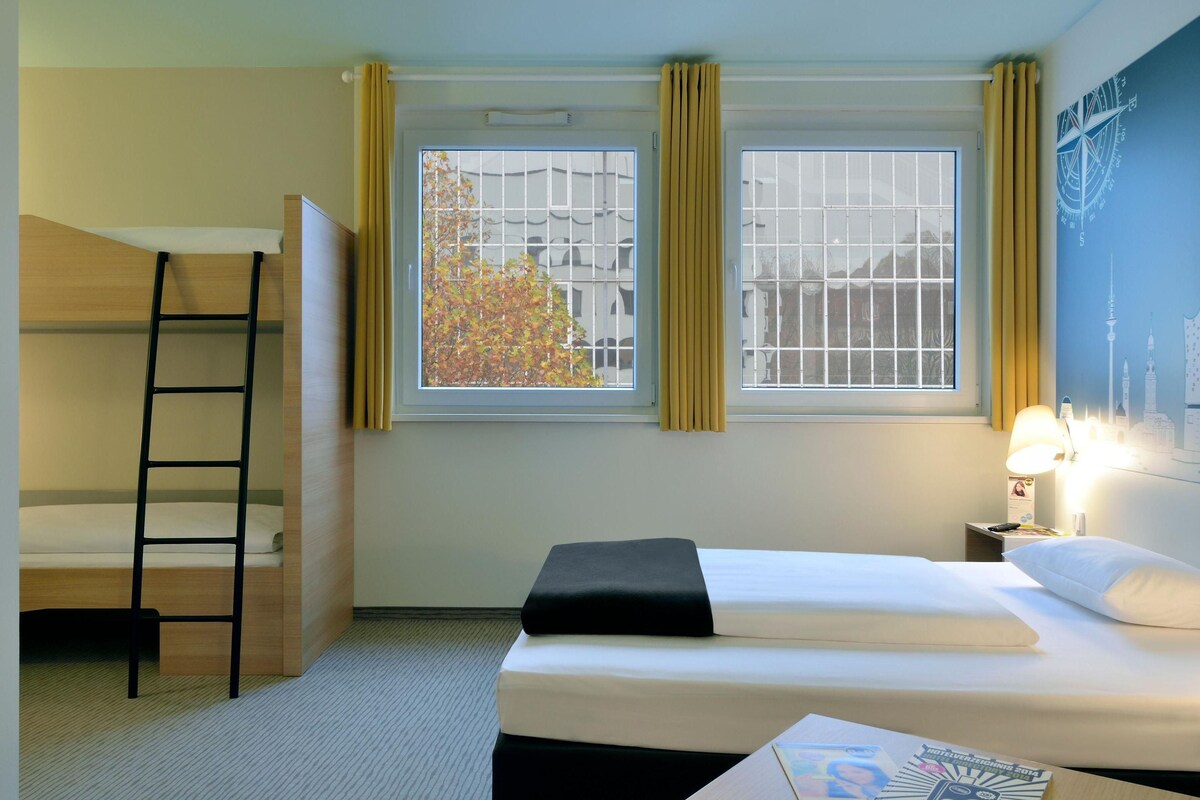 A hotel room with a bunk bed, a single bed, a window with curtains, a nightstand with a lamp, and a wall with a compass design.