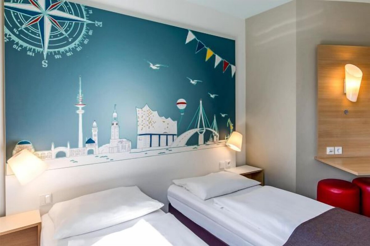 Habitación de hotel con dos camas, un mural de paisaje urbano y cabecera de madera con una lámpara.