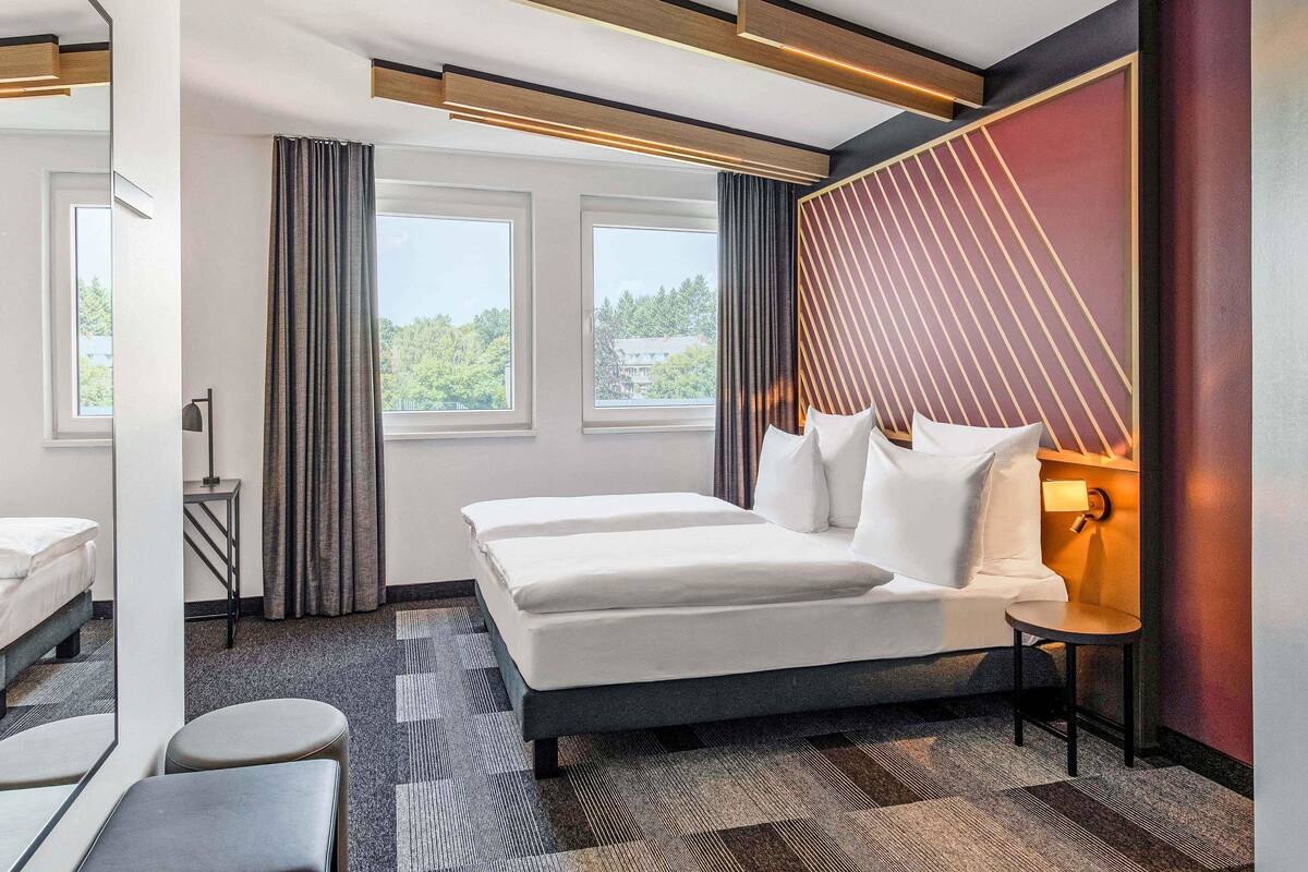 A modern hotel room with a large bed, a bedside table, a mirror, and a view of greenery outside.