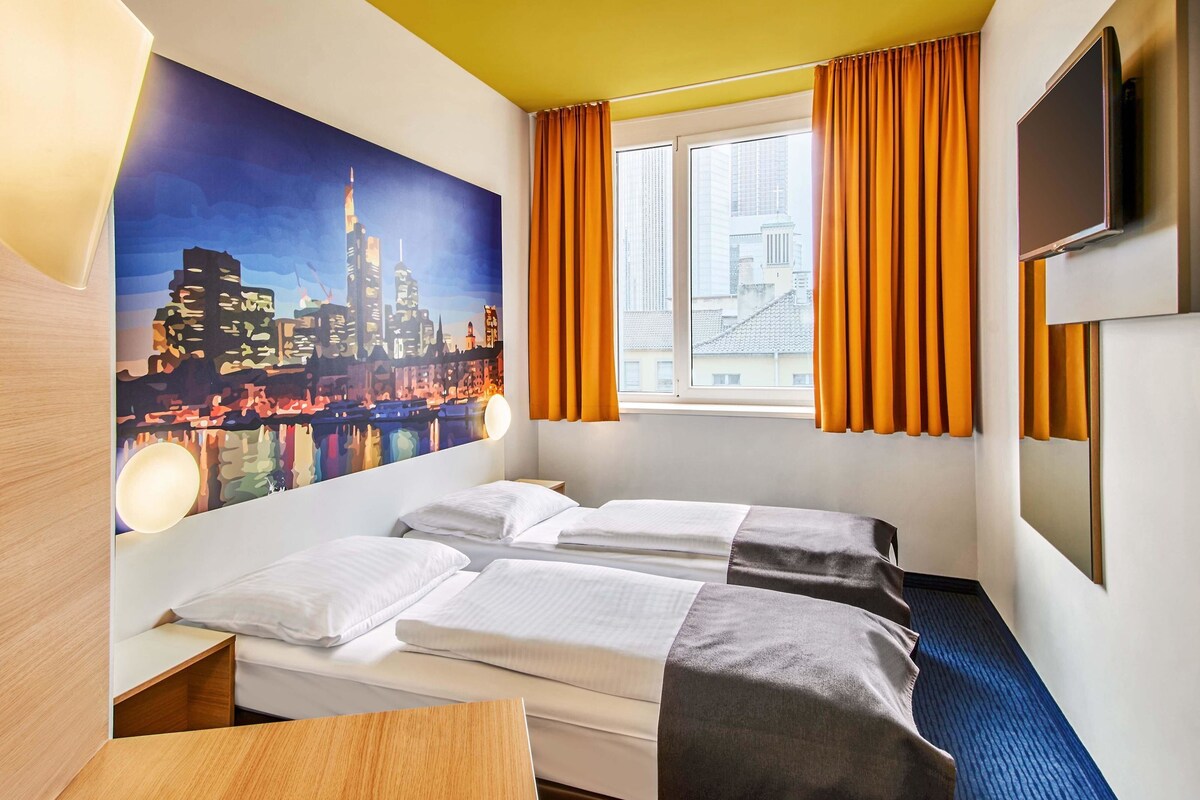 A hotel room with two beds, a large window with orange curtains, a cityscape mural on the wall, and a flat-screen TV mounted on the wall.