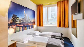 A hotel room with two beds, a large window with orange curtains, a cityscape mural on the wall, and a flat-screen TV mounted on the wall.