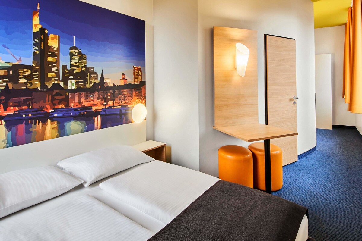 A hotel room with a bed, a nightstand, a mirror, a wall-mounted light, and a cityscape painting.