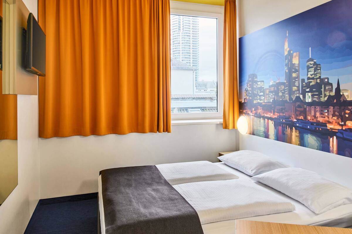 Ein Hotelzimmer mit einem Bett, einem Fenster mit Vorhängen, einem Stadtbildgemälde und einem Flachbildfernseher an der Wand.