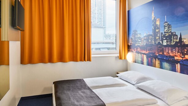 A hotel room with a bed, a window with curtains, a cityscape painting, and a flat-screen TV on the wall.