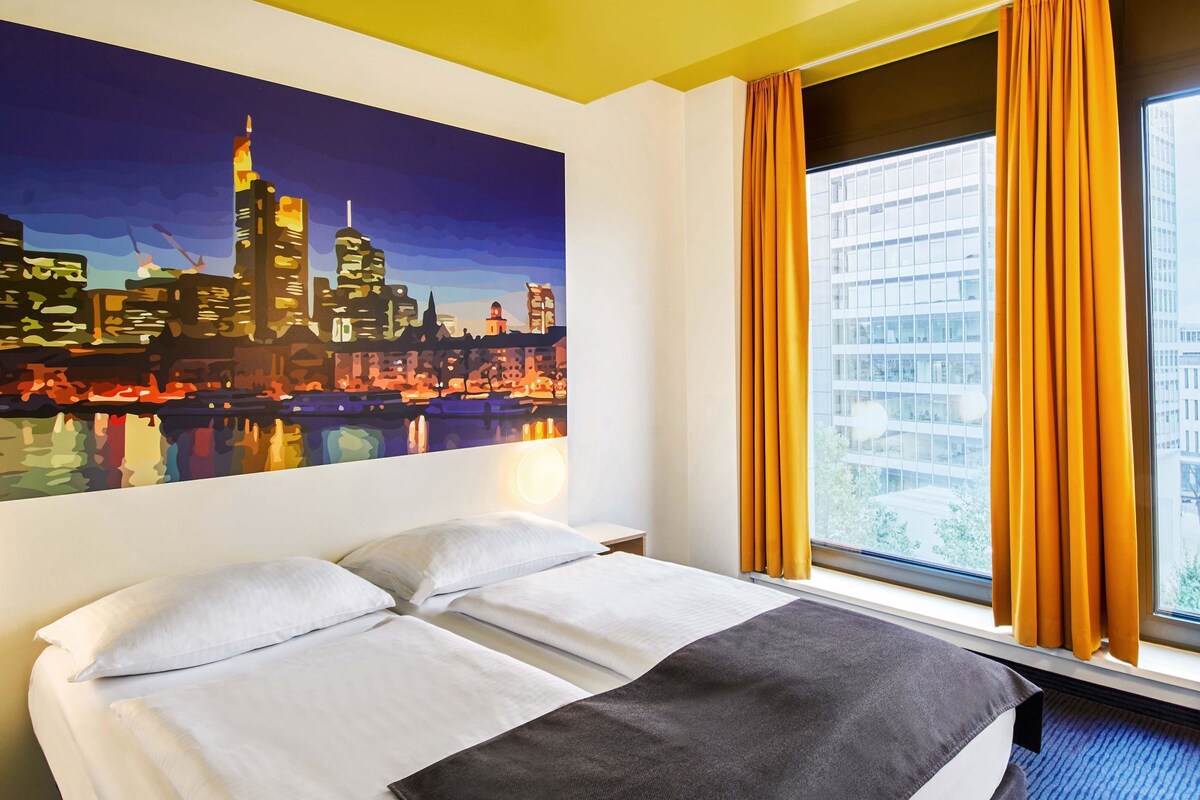 A hotel room with a bed, a cityscape painting, a window with curtains, and a view of buildings.
