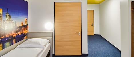 Une chambre d’hôtel avec un lit, une porte en bois, une moquette bleue et un tableau représentant un paysage urbain accroché au mur.