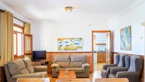 Una sala de estar con un sofá, una mesa de centro y un televisor. Hay un cuadro en la pared y se ve una cocina a través de una puerta abierta.