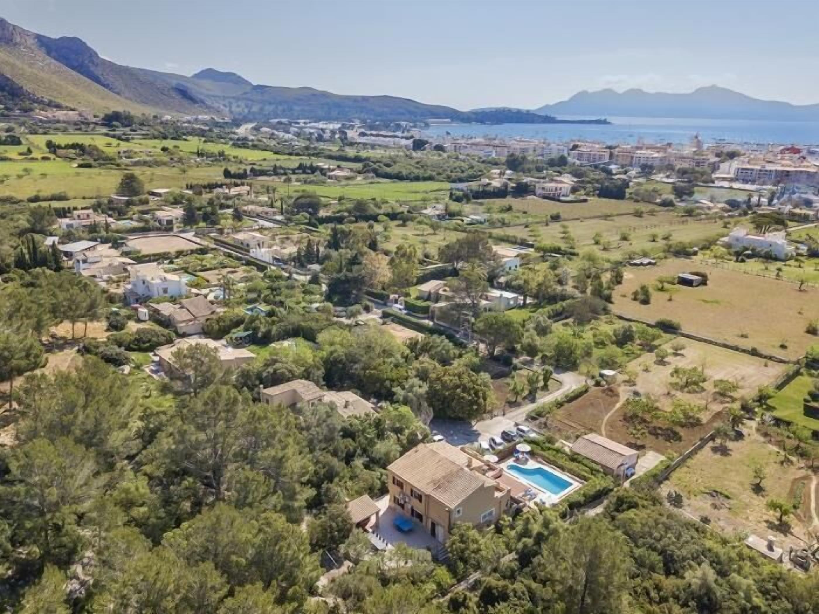 An aerial view of a coastal town with a prominent villa featuring a swimming pool, surrounded by lush greenery and residential buildings.