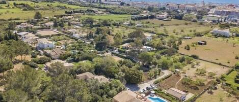 An aerial view of a coastal town with a prominent villa featuring a swimming pool, surrounded by lush greenery and residential buildings.