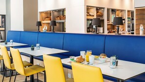 A modern dining area with blue booths and yellow chairs, featuring breakfast items and a view of the kitchen in the background. - B&B Hotel Hamburg City-Ost (Hamburg)
