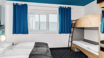 Ein Hotelzimmer mit Etagenbett, einem Einzelbett, einem Fenster mit Vorhängen, einem Schreibtisch und einer Lampe.