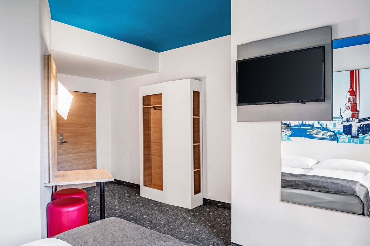 A hotel room with a bed, a TV mounted on the wall, a wooden wardrobe, and a red stool.