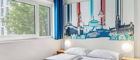 A hotel room with a bed, a window with blue curtains, a cityscape mural on the wall, and a nightstand with a lamp.