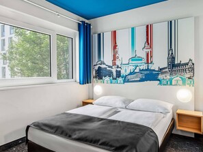 A hotel room with a bed, a window with blue curtains, a cityscape mural on the wall, and a nightstand with a lamp.