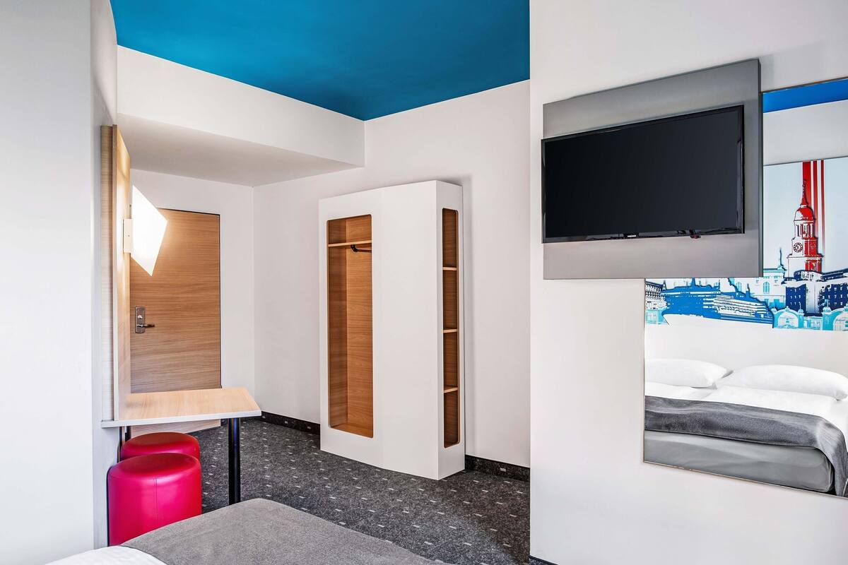 A hotel room with a bed, a TV mounted on the wall, a wooden wardrobe, and a red stool.