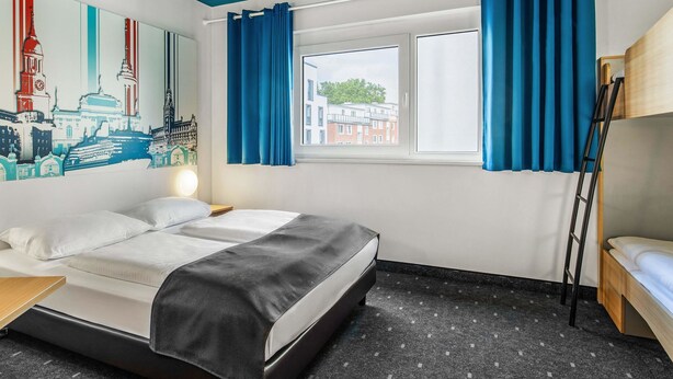 A modern hotel room with a bed, a small table, a window with curtains, a wall with a cityscape mural, and a bunk bed. - B&B Hotel Hamburg City-Ost (Hamburg)