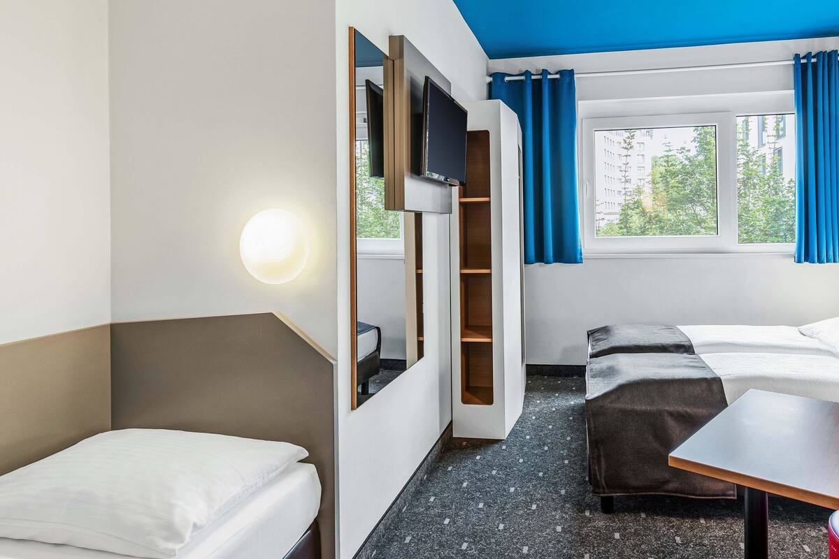 A hotel room with a bed, a desk, a TV mounted on the wall, and a window with curtains.