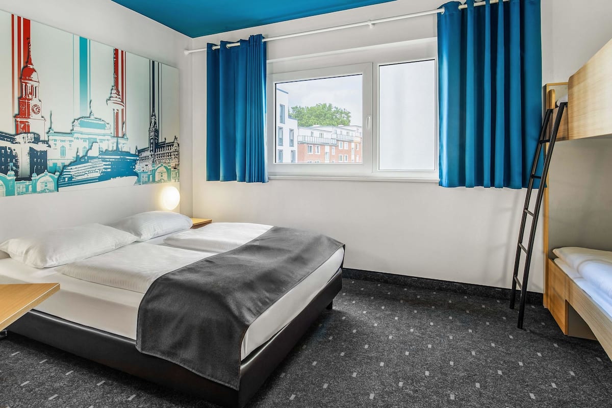 A modern hotel room with a bed, a small table, a window with curtains, a wall with a cityscape mural, and a bunk bed.