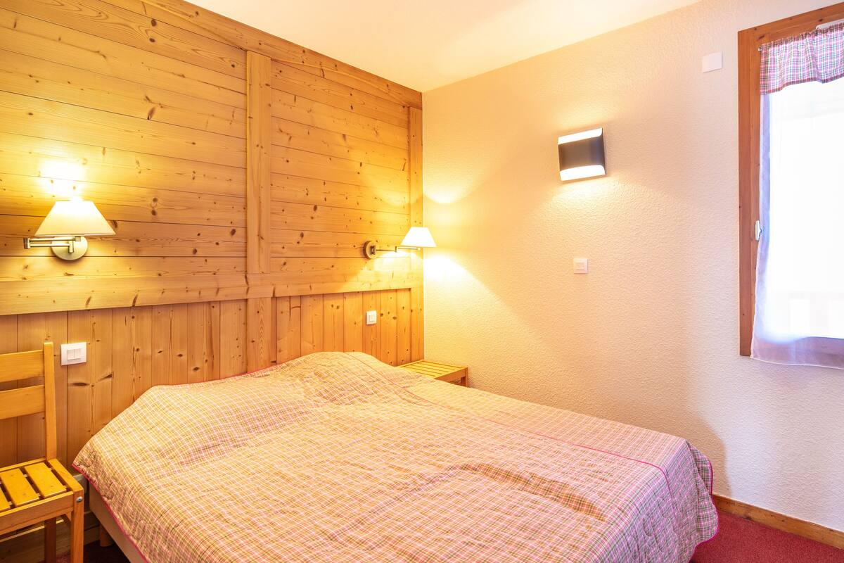 Ein Schlafzimmer mit Holzwandverkleidung, einem Bett mit kariertem Bettbezug, zwei Nachttischlampen, einem Nachttisch aus Holz und einem Fenster mit Vorhang.