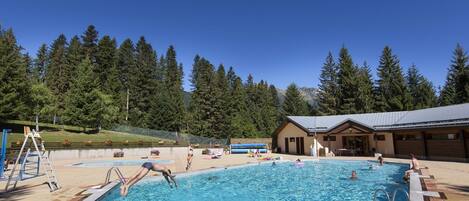 Une piscine sous un ciel bleu azur, entourée d’arbres et bordée d’un bâtiment doté d’un toit en pente.