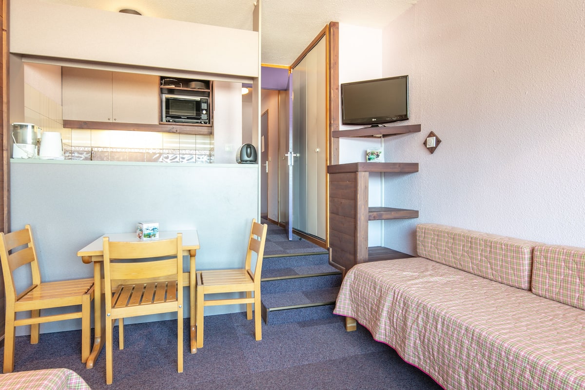 Ein kompaktes Zimmer mit Küchenzeile, einer Essecke mit Tisch und Stühlen, einer Couch und einem an der Wand befestigten Fernseher.