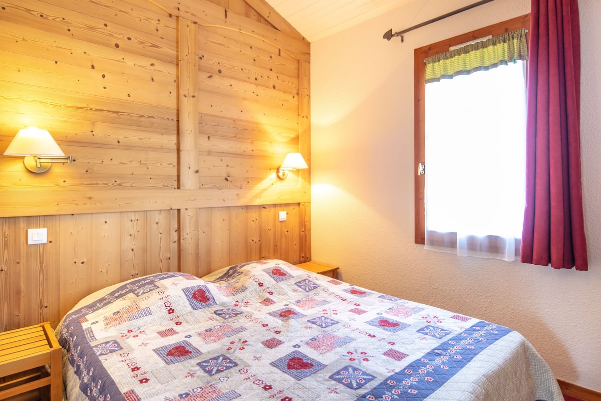 Ein Zimmer mit Holzwänden, einem Bett, zwei Wandleuchten und einem Fenster mit Vorhang.