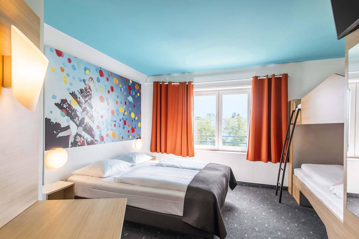 A hotel room with a bed, a desk, a window with curtains, a bunk bed, and a colorful wall decoration.