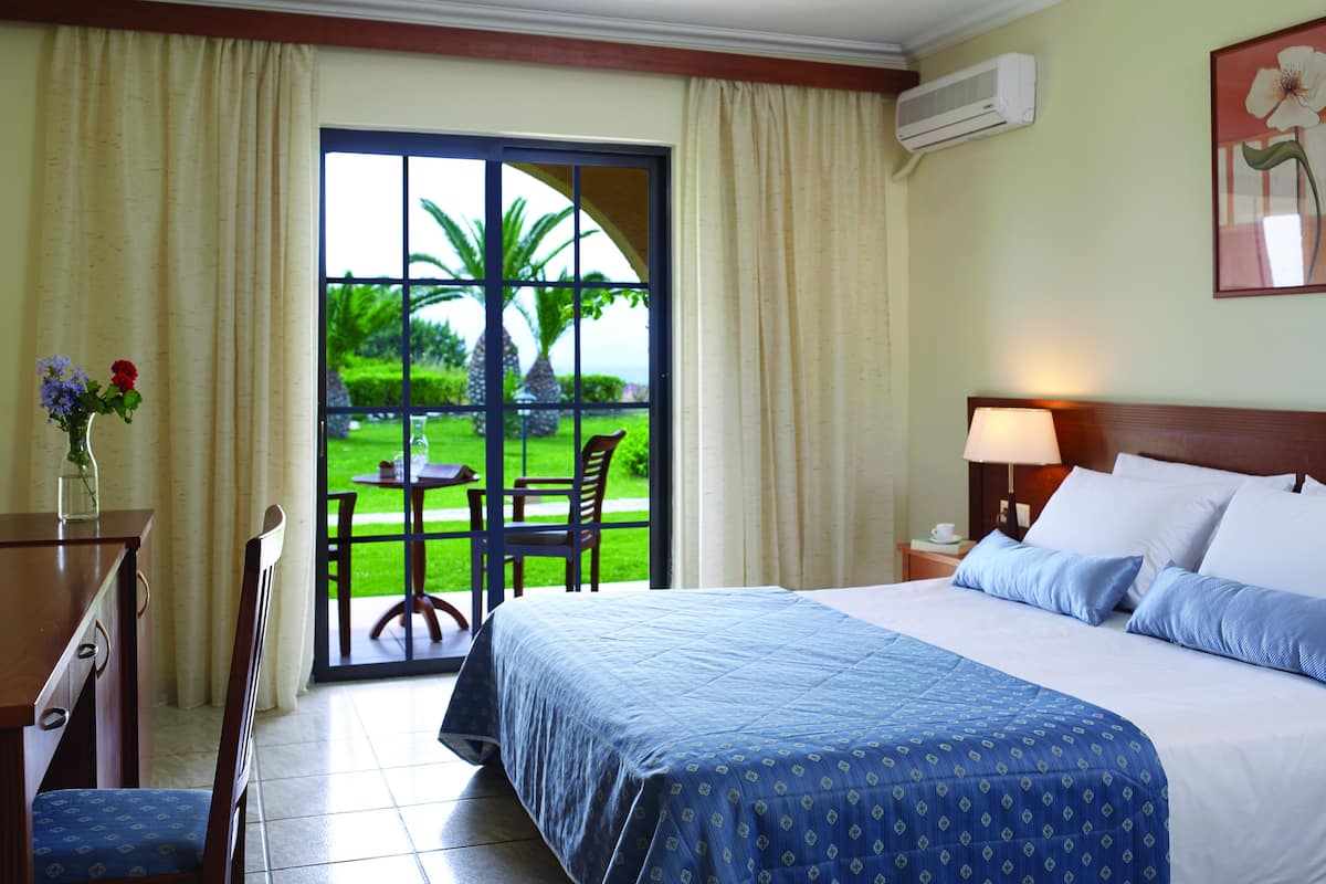A hotel room with a bed, a desk with a vase of flowers, a chair, a table with chairs, and a view of a garden through a glass door.