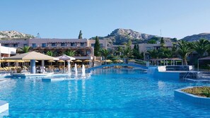 A resort pool area with multiple lounge chairs and umbrellas, a central fountain, and a mountain backdrop.