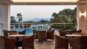 A balcony with wicker chairs and tables overlooking a pool and mountains. - Porto Bello Beach - All Inclusive (Kos)