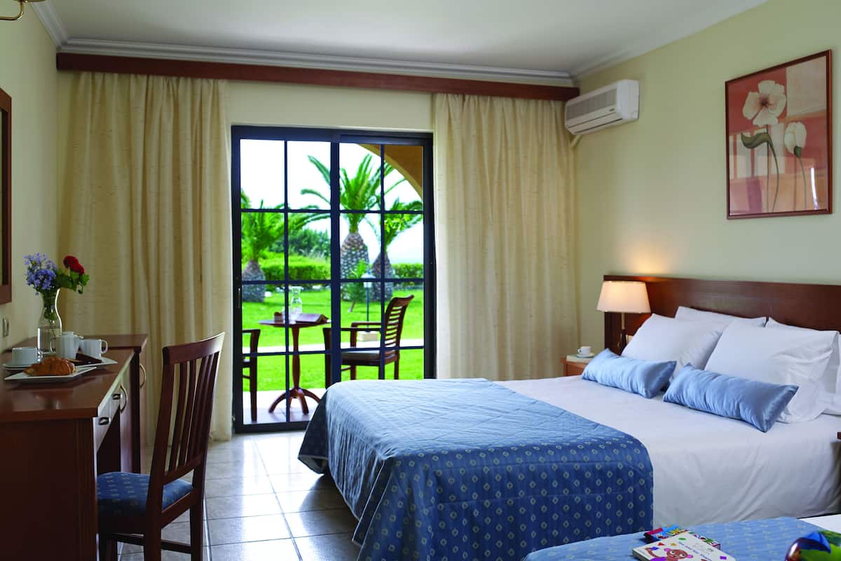 A hotel room with a bed, a desk with a chair, a small table with a vase of flowers, and a view of a garden through a glass door.