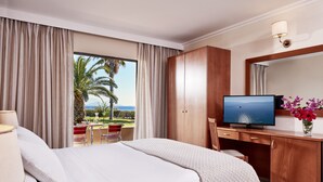 A hotel room with a large bed, a desk with a television, a wardrobe, and a view of palm trees and the ocean. - Porto Bello Beach - All Inclusive (Kos)