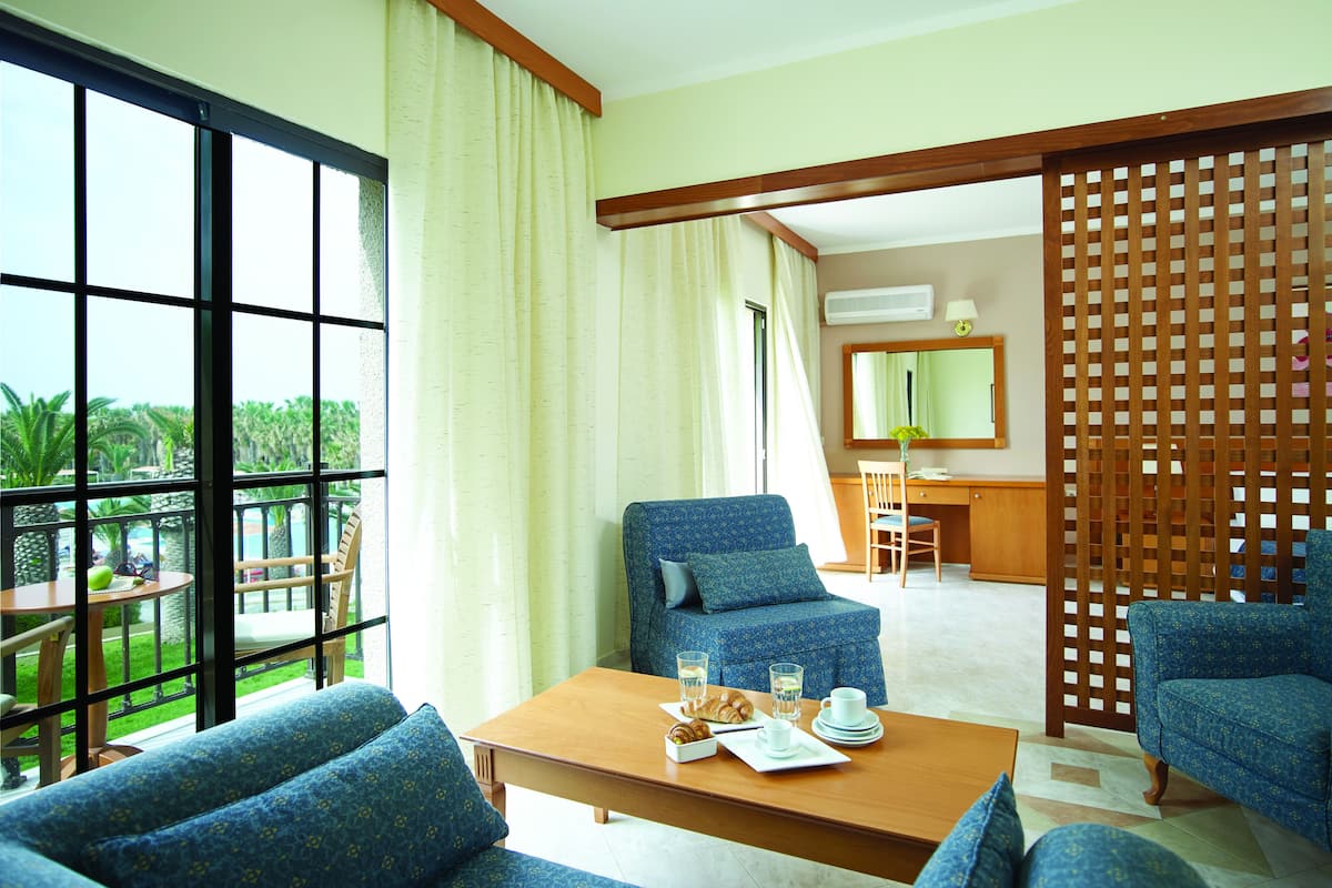 A hotel room with a balcony, a dining area with a table set for two, and a seating area with blue upholstered furniture.