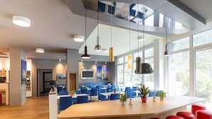 A modern hotel lobby with a large wooden reception desk, red bar stools, and blue chairs. There are large windows providing natural light and a view of the outdoors. - B&B HOTEL Stuttgart-Airport/Messe (Stuttgart)