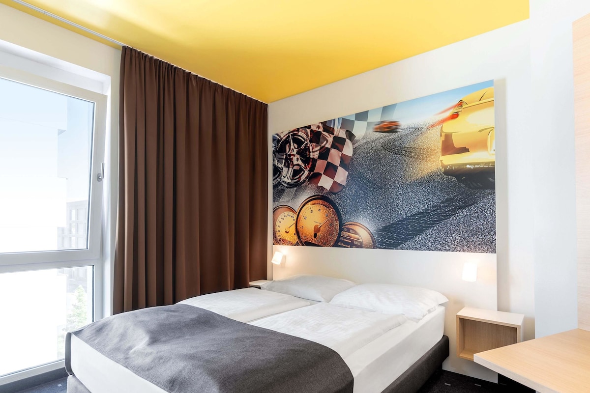 A hotel room with a bed, a desk, a chair, a window with curtains, and a wall with a painting.