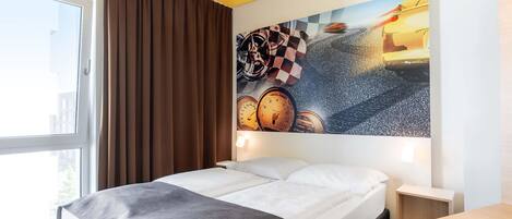 Une chambre d’hôtel avec un lit, un bureau, une chaise, une fenêtre avec des rideaux et un mur orné d’un tableau.
