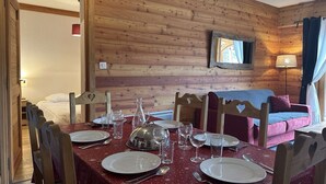 A dining area with a wooden wall, a table set for four, and a sofa in the background. - Ski-In/Ski-Out Apartment, 6 Guests, Terrace & Private Ensuite (Montgenèvre)