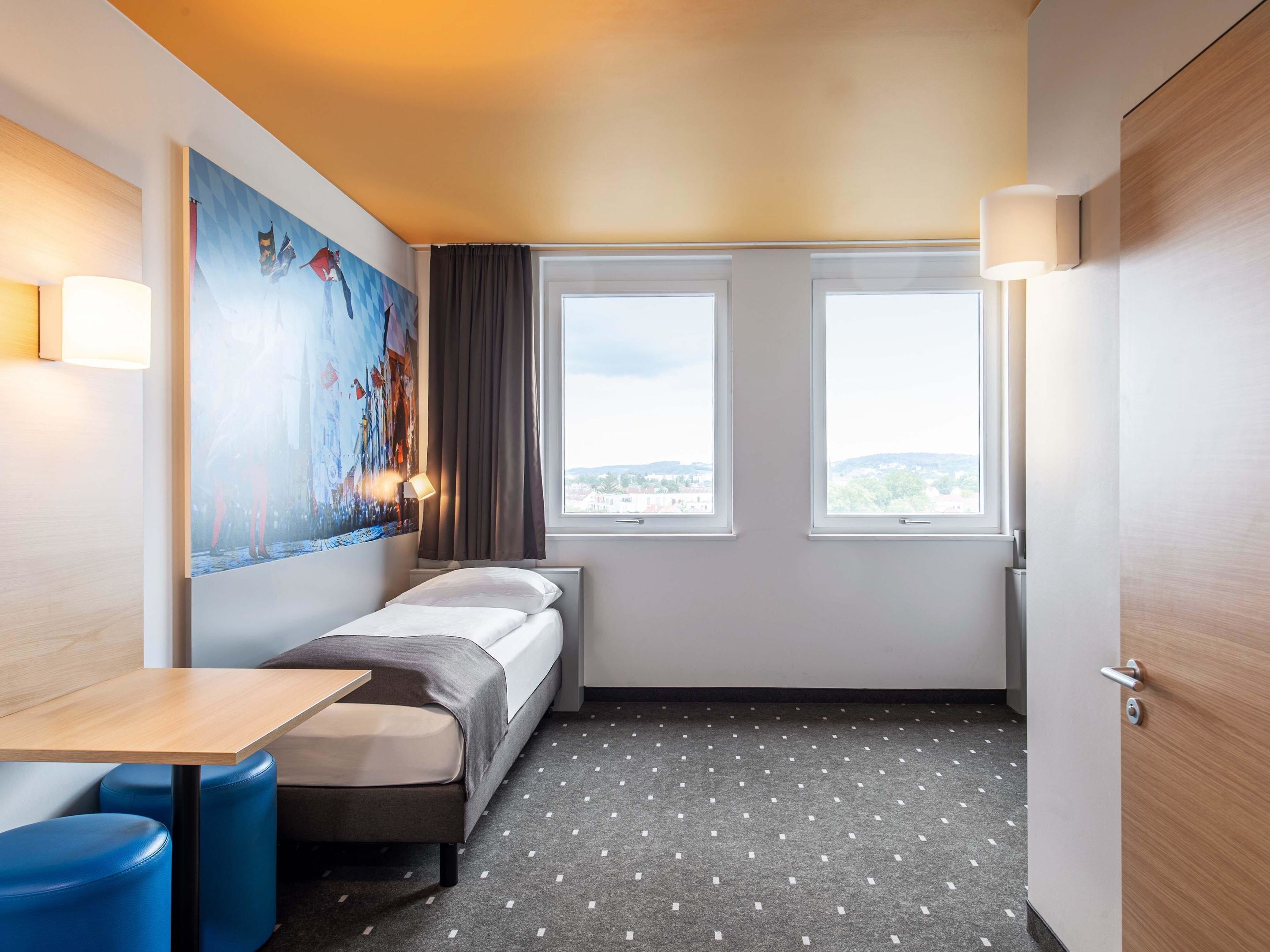 A small hotel room with a bed, a table, two windows, a painting on the wall, and a door.