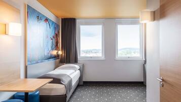 A small hotel room with a bed, a table, two windows, a painting on the wall, and a door.