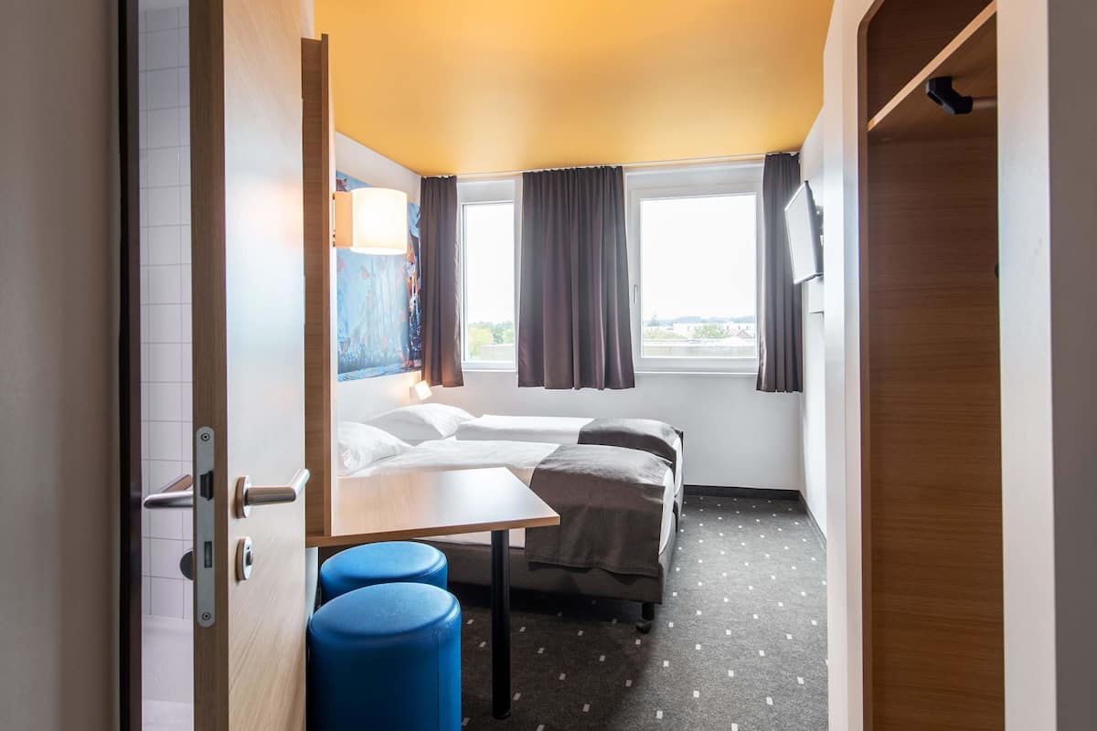 A hotel room with a bed, a desk, two blue stools, a window with curtains, and a painting on the wall.