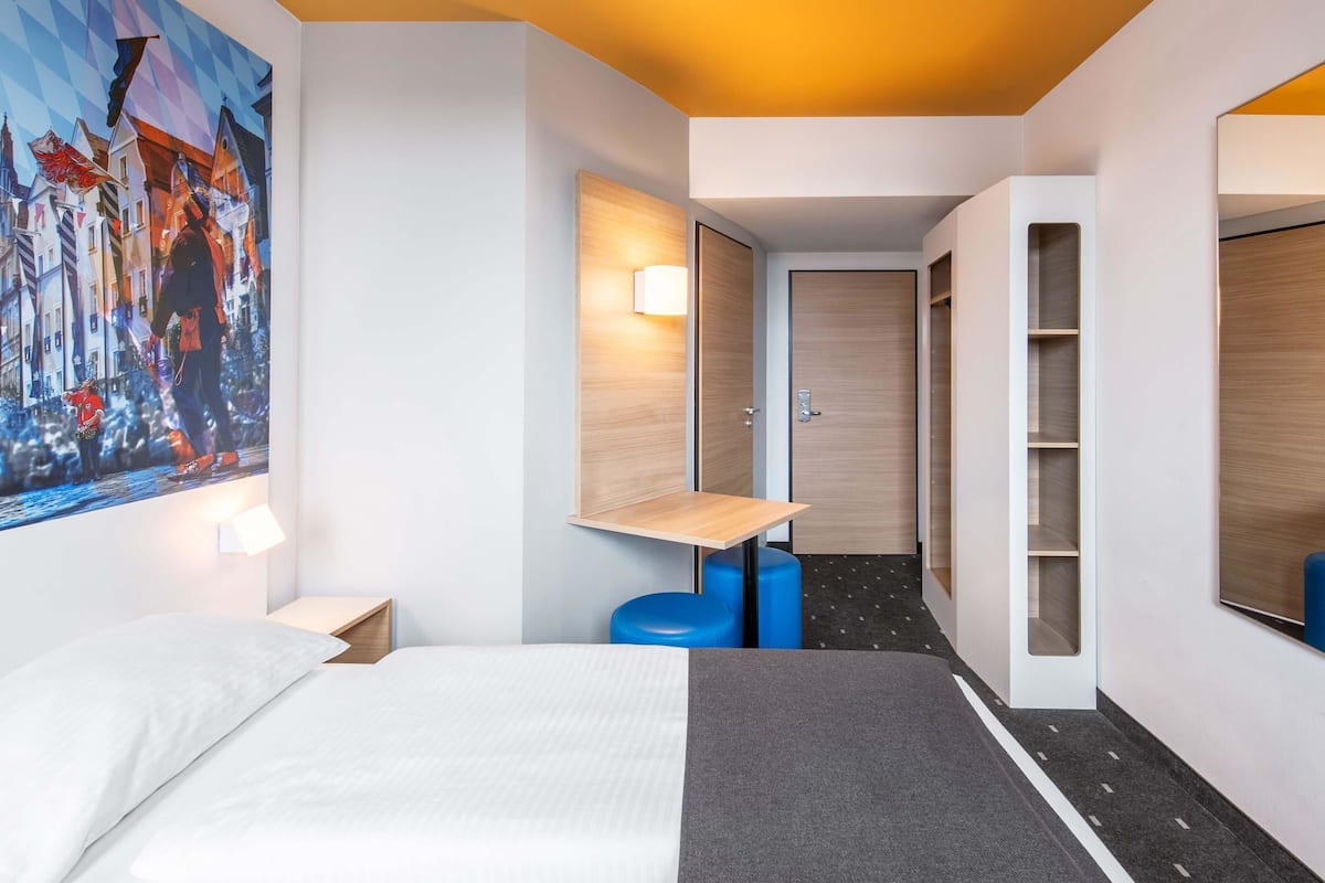 A hotel room with a bed, a desk, a chair, a wardrobe, and a painting on the wall.