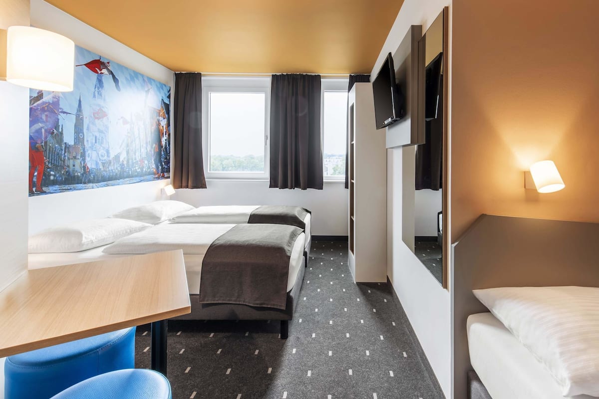A hotel room with two beds, a desk, a TV, and a painting of a cityscape.
