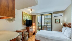 A hotel room with a bed, a desk, a television, a balcony with a view of buildings, and an air conditioning unit.
