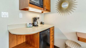 A compact kitchenette with a microwave, coffee maker, and a small refrigerator. There is a wooden cabinet above the counter and a decorative sunburst mirror on the wall.