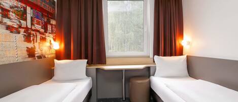 Una habitación pequeña y funcional con dos camas individuales, un escritorio pequeño y una ventana con cortinas.