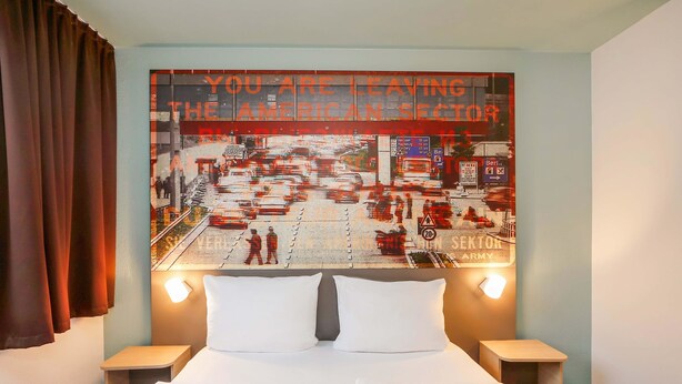 Een hotelkamer met een bed, twee nachtkastjes en een groot muurschildering waarop een stadsstraat te zien is, met een boodschap over vertrek uit het Amerikaanse deel.