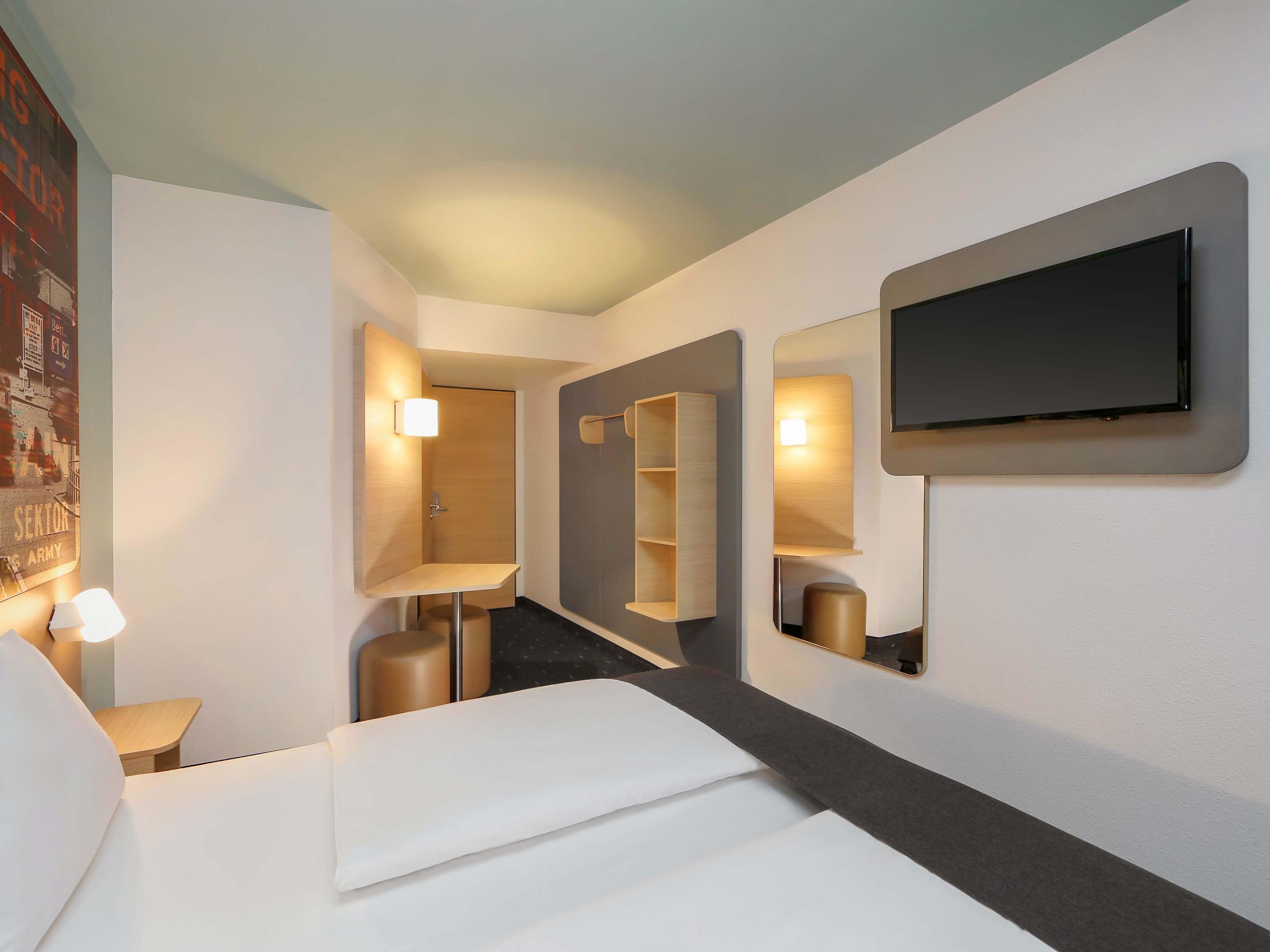 A modern hotel room with a bed, a TV mounted on the wall, and a mirror reflecting the room.