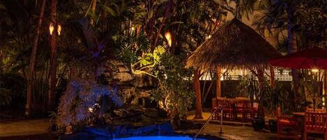 Un espace piscine nocturne doté d’une structure à toit de chaume et de chaises rouges, le tout entouré d’une végétation tropicale luxuriante.